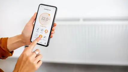 Eine Person bedient ein Smartphone mit einer Temperatur-App vor einem weißen Heizkörper. Die Temperaturanzeige beträgt 22 Grad Celsius.