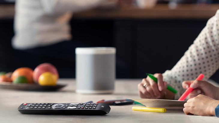 Auf einem Tisch liegen eine Fernbedienung, Obst, ein grauer Lautsprecher und Personen, die mit Stiften malen.