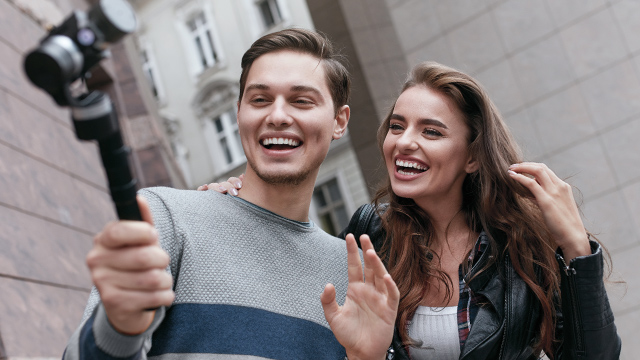 Ein Mann hält eine Kamera, während er und eine Frau lachen; beide stehen vor einem Gebäude.