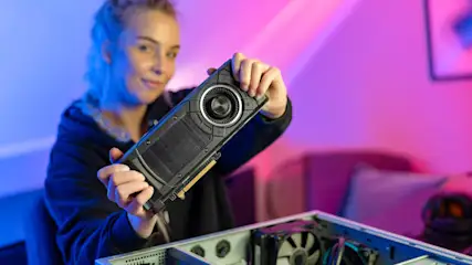 Eine Person hält eine Grafikkarte über einem offenen Computergehäuse vor einem violett-blauen Hintergrund.