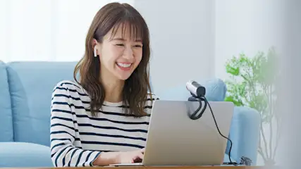 Eine lächelnde Person mit Kopfhörern benutzt einen Laptop mit Webcam. Im Hintergrund ein blaues Sofa und eine Zimmerpflanze.