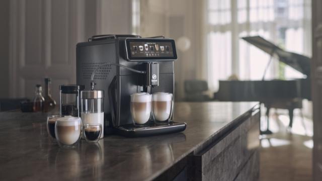Kaffeemaschine mit Tassen auf einer Theke, Gläser und Flaschen daneben. Im Hintergrund ist ein Klavier vor einem Fenster zu sehen.