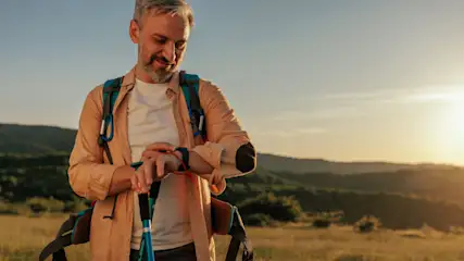Ein Mann mit Rucksack und Wanderstöcken kontrolliert seine Uhr, im Hintergrund Hügel und ein heller Himmel.