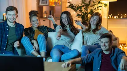 Fünf Personen sitzen auf einer Couch und lächeln. Sie tragen Jeans und Freizeitkleidung. Im Hintergrund sind ein Fernseher und ein Regal zu sehen.