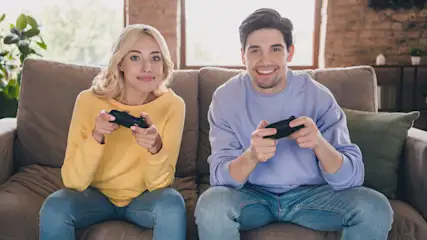 Ein Paar sitzt auf einem Sofa und hält Controller. Die Frau trägt einen gelben Pullover, der Mann einen lila Pullover und Jeans.