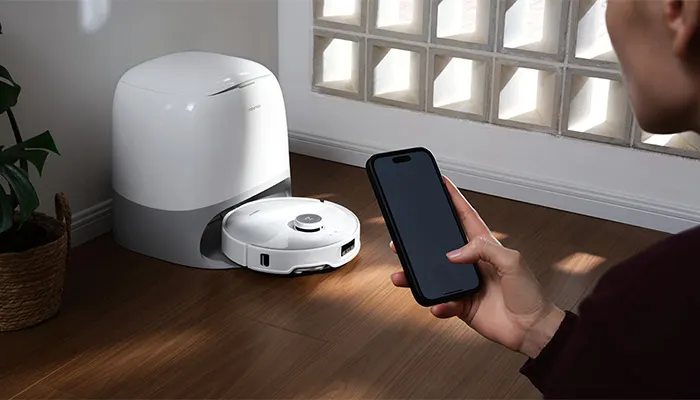 Ein weißer Roboterstaubsauger steht neben einer Dockingstation auf einem Holzboden; eine Person hält ein Smartphone.