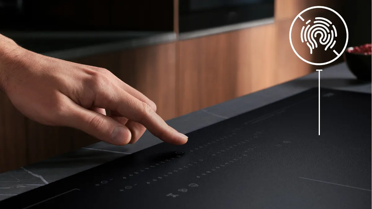 Eine Hand berührt ein schwarzes Kochfeld mit Touchscreen-Bedienfeld. Im Hintergrund ist eine hölzerne Küchenzeile sichtbar.