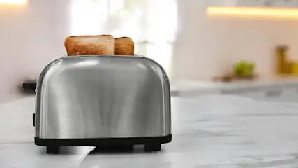 Auf einer hellen Arbeitsfläche steht ein silberner Toaster mit zwei Scheiben Toastbrot. Im Hintergrund ist eine Küche zu sehen.