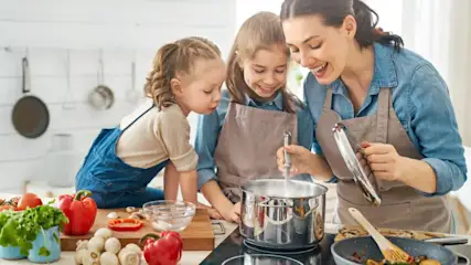 In einer Küche kocht eine Frau mit zwei Kindern. Auf dem Tisch stehen Gemüse, Pilze und Kochutensilien.