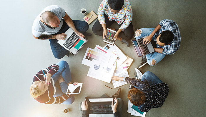 Blick von oben auf eine Arbeitsgruppe. Junge Menschen sitzen am Boden und halten ihren Laptop oder ein Tablet in der Hand.