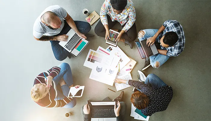 Blick von oben auf eine Arbeitsgruppe. Junge Menschen sitzen am Boden und halten ihren Laptop oder ein Tablet in der Hand.