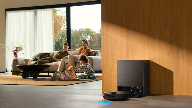  Saugroboter mit Absaugstation in einem modernen Wohnzimmer. Während der Roboter den Boden reinigt, entspannen zwei Erwachsene und ein Kind auf dem Sofa im Hintergrund.
