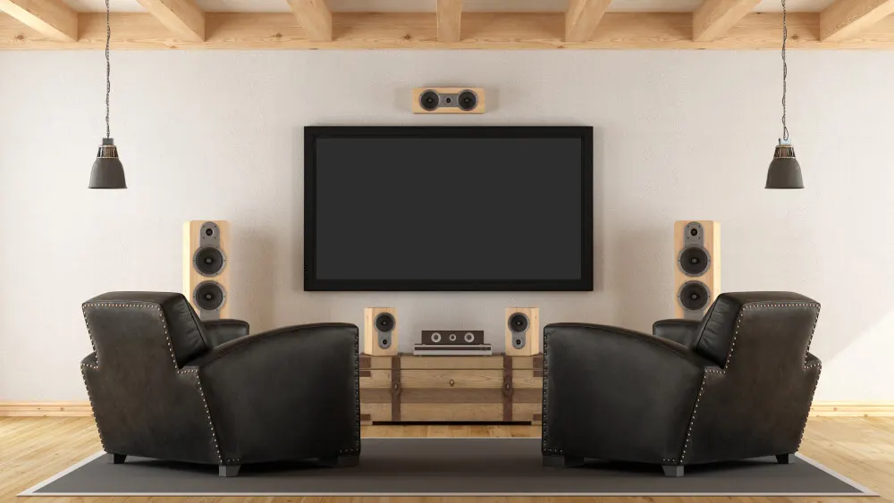 Ein Wohnzimmer mit zwei Sesseln, einem Fernseher, Lautsprechern und einer Kommode. Deckenbalken und zwei Hängelampen sind auch zu sehen.