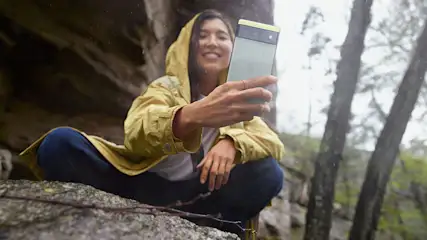 Eine Person mit gelber Jacke und blauem Hosen hockt auf einem Felsen und hält ein Handy. Bäume sind im Hintergrund.