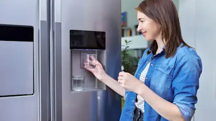 Frau mit blauem Hemd füllt ein Glas Wasser an einem silbernen Kühlschrank mit Wasserspender.