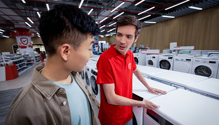 In einem Geschäft stehen zwei Männer vor einer Reihe von Waschmaschinen. Einer trägt ein rotes Hemd, der andere eine Jacke.