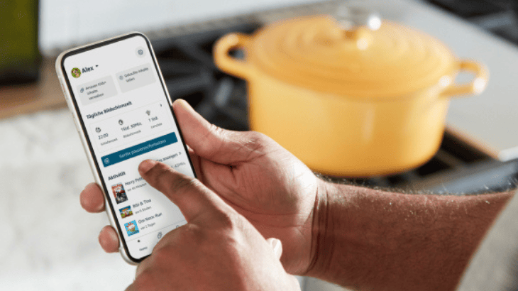 Mann hält ein Smartphone in seiner Hand, im Hintergrund steht ein gelber Kochtopf auf einem Herd. Das Smartphone zeigt das Eltern Dashboard von Alexa.