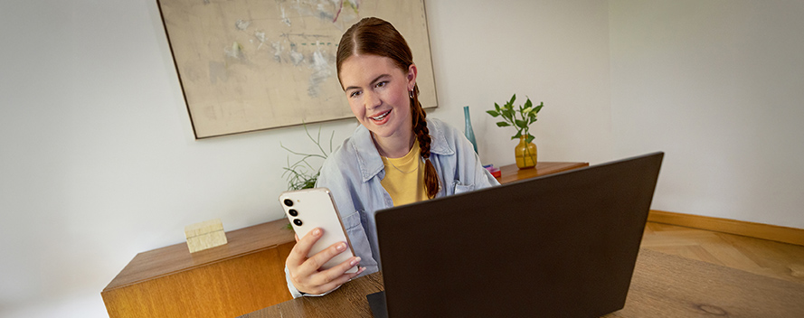 Eine Frau sitzt am Schreibtisch mit Laptop und Handy. Im Hintergrund sind eine Kommode, ein Bild und eine Pflanze.