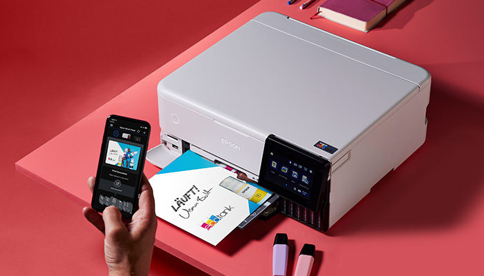 Ein Drucker steht auf einem roten Tisch. Eine Hand hält ein Smartphone. Papier wird bedruckt. Textmarker liegen daneben.