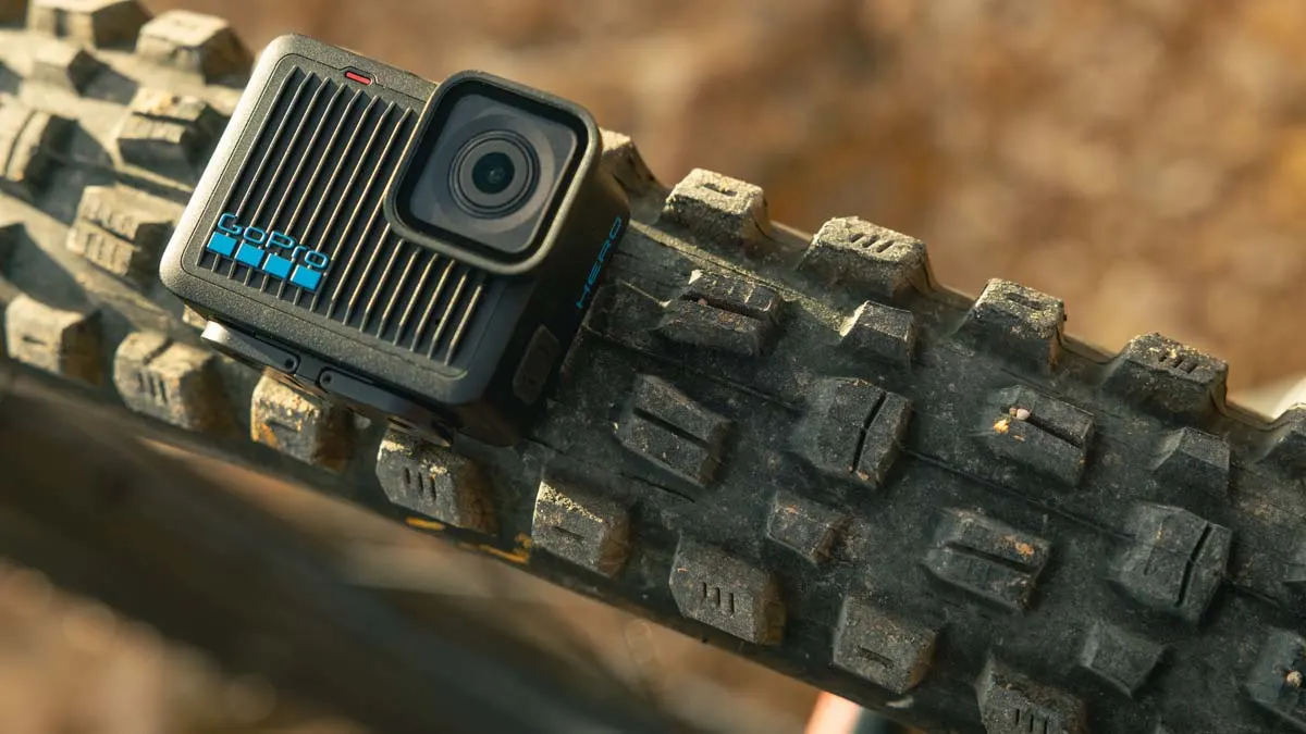 Eine schwarze GoPro-Kamera mit blauem Logo liegt auf einem schmutzigen, schwarzen Fahrradreifen vor einem unscharfen, braunen Hintergrund.