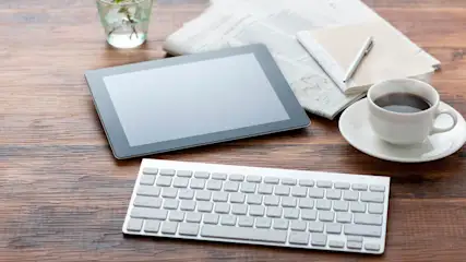 Auf einem Holztisch liegen ein Tablet, eine weiße Tastatur, eine Tasse Kaffee, eine Zeitung, ein Notizblock, ein Stift und ein Glas mit Pflanzen.