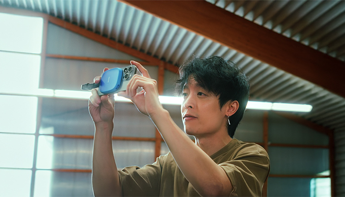 Person hält ein kleines blaues tragbares Gerät mit beiden Händen hoch; Innenraum einer lichtdurchfluteten Halle mit Rippenblech-Decke und Holzbalken. 