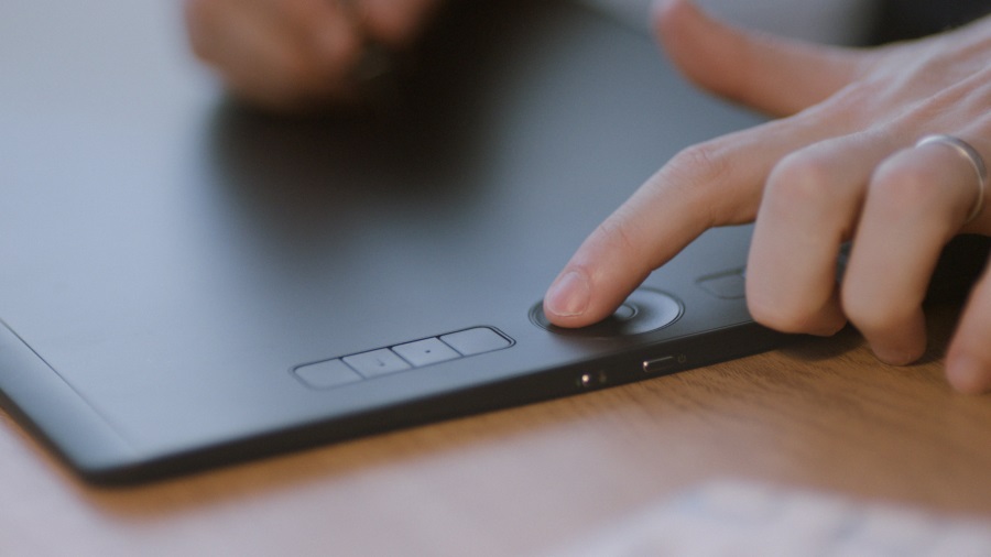 Eine Hand mit einem Ring drückt einen runden Knopf auf einem schwarzen Grafiktablett mit vier quadratischen Tasten.