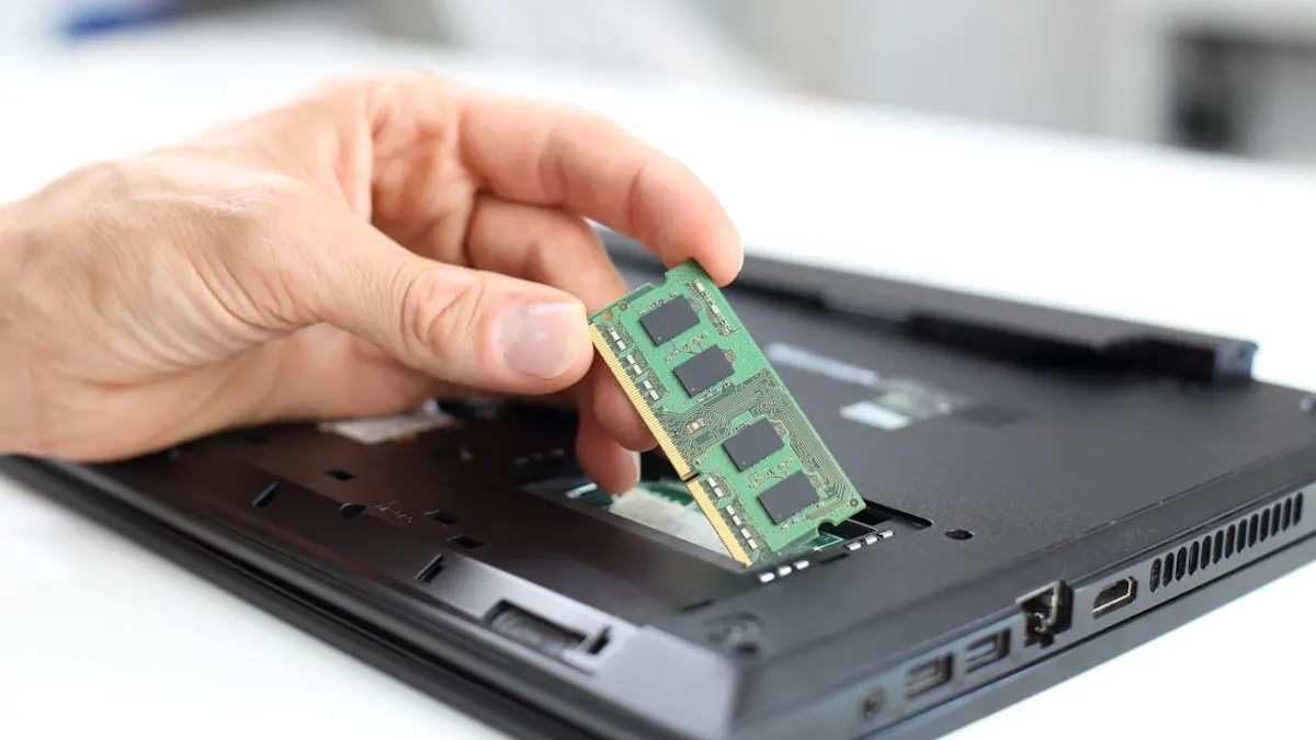 Eine Hand hält einen grünen RAM-Speicher über einen offenen Laptop. Der Laptop ist schwarz und liegt auf einem weißen Tisch.