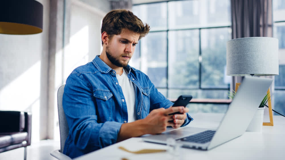 Ein Mann mit einem Smartphone in der Hand sitzt neben einem offenem Laptop.