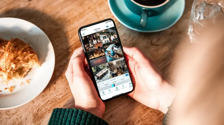 Eine Person hält ein Smartphone über einem Holztisch. Darauf sind ein Croissant, eine Kaffeetasse und ein Glas Wasser zu sehen.