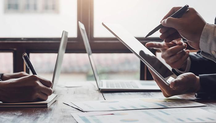 Business-Meeting: Hände schreiben und zeigen auf Tablet; Laptop und Berichte mit Diagrammen auf Holztisch vor Fenster. 
