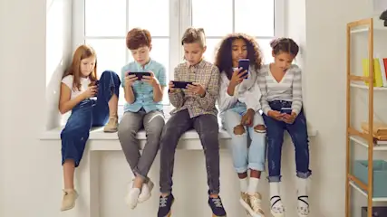 Fünf Kinder sitzen auf einem Fensterbrett und schauen auf ihre Smartphones. Im Hintergrund sind ein Fenster und ein Regal zu sehen.