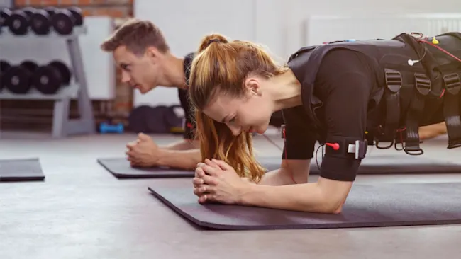 Zwei Personen in schwarzer Sportkleidung machen Planks auf Matten in einem Fitnessstudio mit Gewichten im Hintergrund.