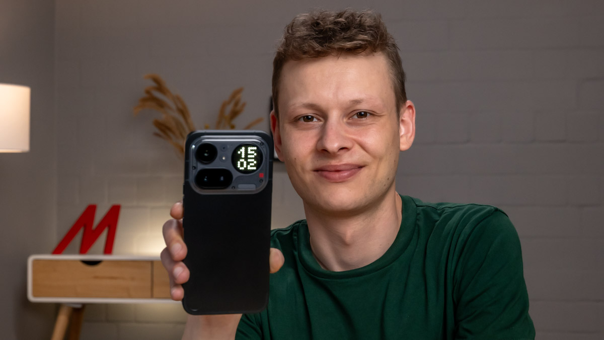 Alex hält das Nothing Phone (4a) Pro mit der Rückseite zur Kamera. Im rechten oberen Eck des Smartphones erkennst du die Uhrzeit auf der Glyph-Matrix.