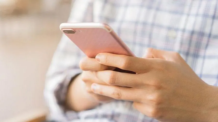 Eine Person hält ein rosafarbenes Smartphone in beiden Händen; kariertes Hemd im Hintergrund.