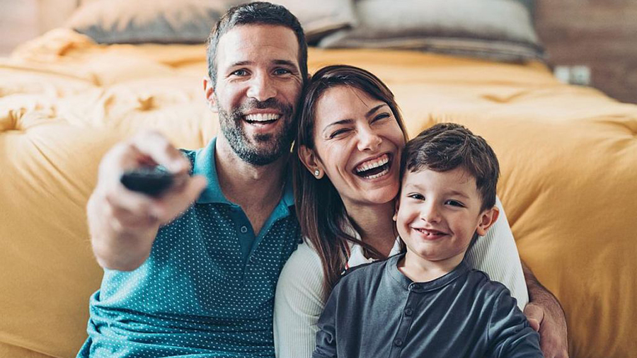 Eine Familie sitzt vor einem gelben Bett. Der Vater hält eine schwarze Fernbedienung. Alle lächeln.
