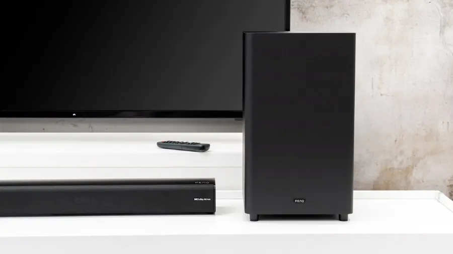 Schwarzer Fernseher, Soundbar, Subwoofer und Fernbedienung auf einem weißen Tisch vor einer Wand.