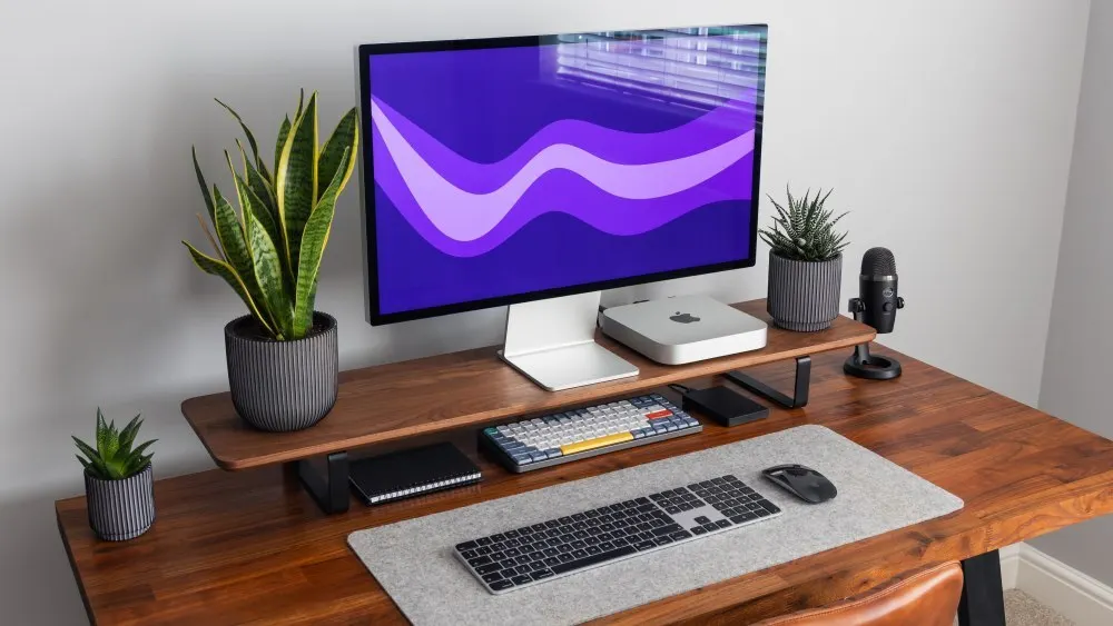 Ein Schreibtisch aus Holz mit einem Monitor, Tastaturen, Maus, Pflanzen und einem Mikrofon vor einer Wand.