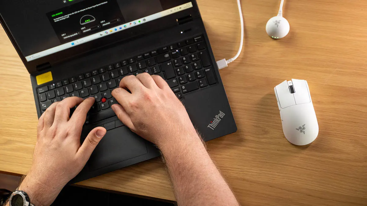 Die weiße Razer Viper V4 Pro liegt rechts neben einem schwarzen ThinkPad-Laptop, auf dem Hände tippen.