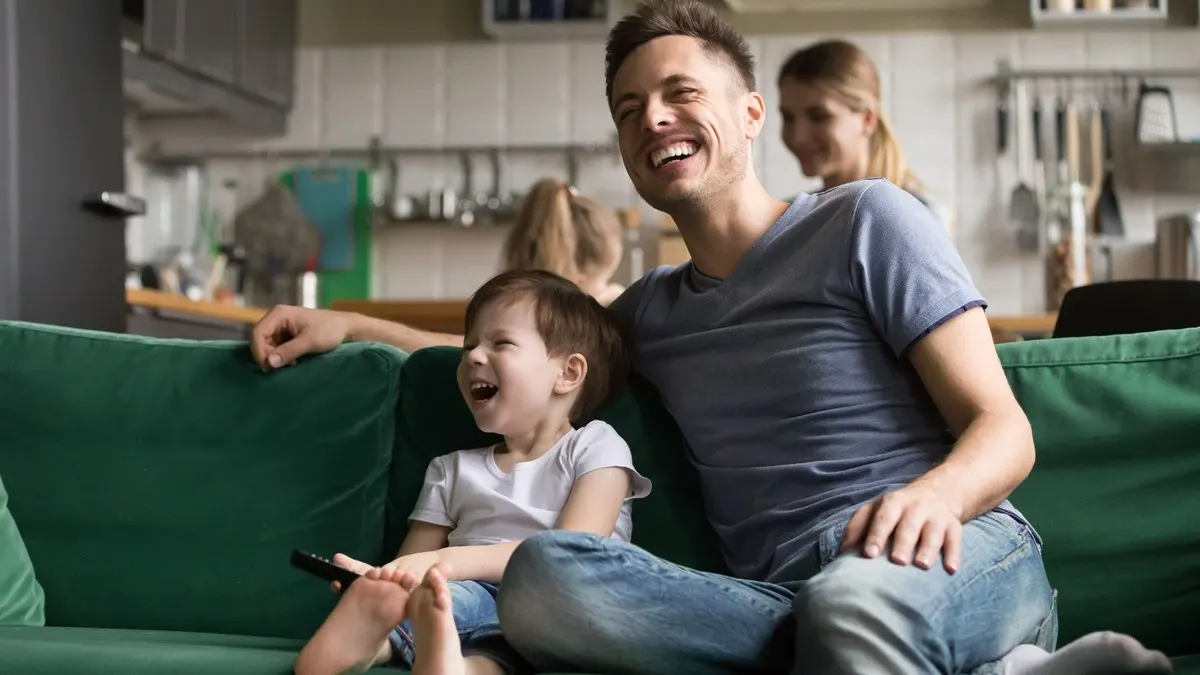 Ein Mann und ein kleiner Junge sitzen auf einem Sofa, das Kind hält eine Fernbedienung in der Hand. Beide lachen.