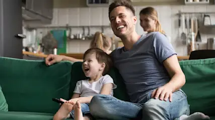 Ein Mann und ein kleiner Junge sitzen auf einem Sofa, das Kind hält eine Fernbedienung in der Hand. Beide lachen.