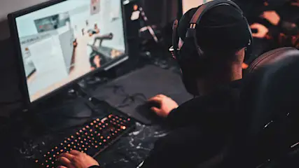 Ein Mann mit Headset und Mütze tippt auf eine beleuchtete Tastatur vor einem Monitor und spielt darauf ein Spiel.
