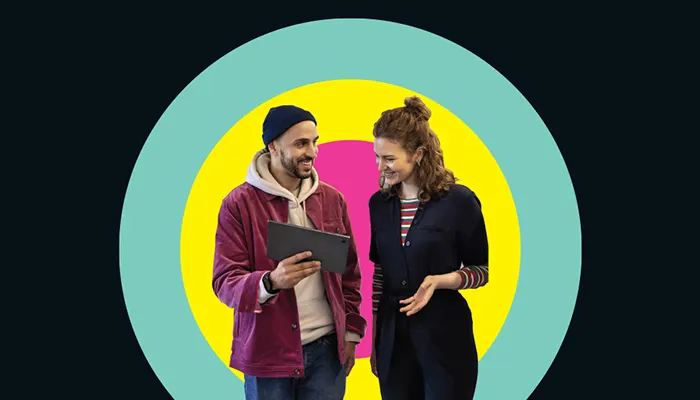 Ein junger Mann mit Mütze und schwarzem Tablet in der Hand im Dialog mit einer jungen Frau vor einem buntgestreiften Bullseye-Hintergrund.