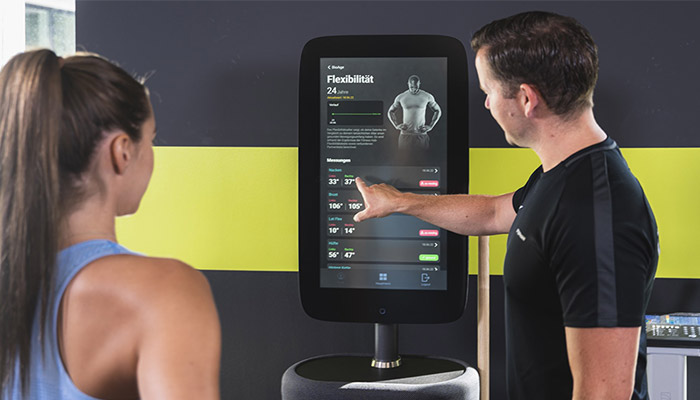 Zwei Personen, die einen großen schwarzen Touchscreen mit Text und Grafiken in einem Fitnessstudio betrachten