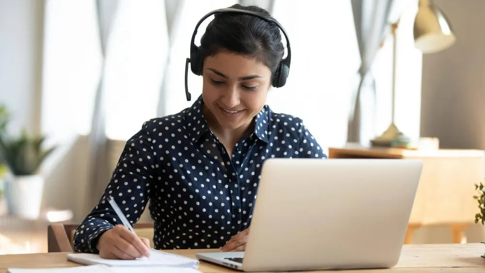 Eine Frau mit Kopfhörern sitzt an einem Tisch mit einem Laptop und schreibt auf Papier.