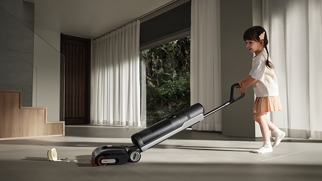 Ein Kind schiebt einen schwarzen Staubsauger auf einem grauen Teppich. Im Hintergrund sind Vorhänge und eine Tür sichtbar.