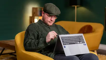 Ein Mann mit Mütze und Brille sitzt in einem gelben Sessel und bedient einen silbernen Laptop. Im Hintergrund sind ein grüner Wand und ein gelbes Sofa.