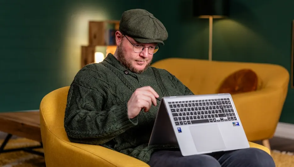 Ein Mann mit Mütze und Brille sitzt in einem gelben Sessel und bedient einen silbernen Laptop. Im Hintergrund sind ein grüner Wand und ein gelbes Sofa.