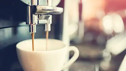 Eine Kaffeemaschine füllt eine weiße Tasse mit braunem Kaffee; silberne Maschine und verschwommener Hintergrund.