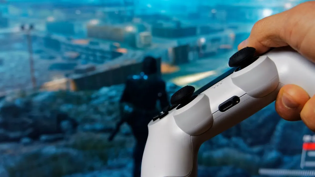 Eine Hand hält einen weißen Controller mit schwarzen Joysticks vor einem verschwommenen Bildschirm. Auf dem Bildschirm befindet sich eine Figur.